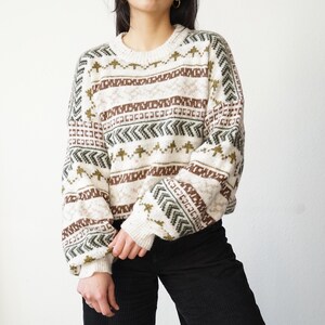 Könnte beinhalten: Cremefarbener Pullover mit geometrischem Muster in Braun, Grün und Weiß. Der Pullover hat einen Rundhalsausschnitt und lange Ärmel mit gerippten Bündchen. Das Design zeigt sich wiederholende Formen und Linien.