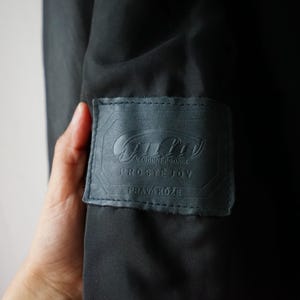 May include: Close-up of a dark gray leather label with embossed text. The label is rectangular with rounded corners and is sewn onto a dark fabric. The text includes the words "NARODNI PODNIK", "PROSTEJOV", and "PRAVA KUZE".