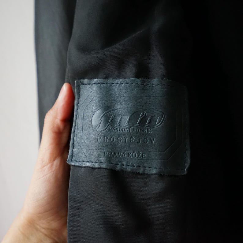 May include: Close-up of a dark gray leather label with embossed text. The label is rectangular with rounded corners and is sewn onto a dark fabric. The text includes the words "NARODNI PODNIK", "PROSTEJOV", and "PRAVA KUZE".