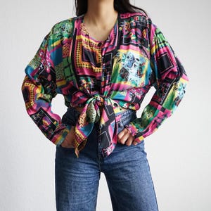 Könnte beinhalten: Eine bunte Langarmbluse mit einem lebendigen geometrischen Muster in Pink, Schwarz, Grün und Blau. Die Bluse ist an der Taille gebunden und wird mit einer blauen Jeans kombiniert. Das Oberteil hat einen V-Ausschnitt und Knöpfe.