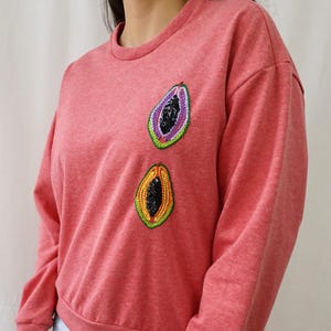 Puede incluir: Una sudadera de color coral con dos diseños bordados de fruta de papaya. Las rodajas de papaya presentan centros de lentejuelas negras, rodeados de costuras coloridas. La sudadera tiene cuello redondo y mangas largas.
