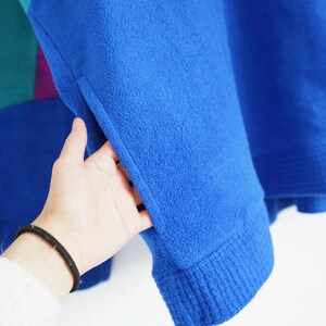 Op de afbeelding: Een close-up van een blauwe fleece trui met een geribde manchet. De trui heeft een zak en kleurgeblokte secties in teal en paars. De mouw wordt door een hand opengehouden.