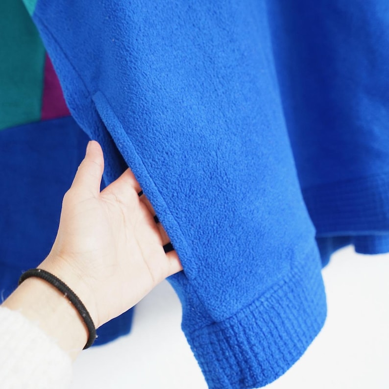 Op de afbeelding: Een close-up van een blauwe fleece trui met een geribde manchet. De trui heeft een zak en kleurgeblokte secties in teal en paars. De mouw wordt door een hand opengehouden.