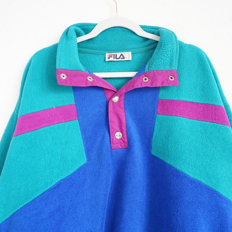 Op de afbeelding: Een vintage Fila fleece trui in teal, blauw en paars. Het colorblock-ontwerp heeft een teal body met paarse accenten en een blauw middenpaneel. De kraag en sluiting hebben drukknoopsluitingen en het Fila-logo is zichtbaar.