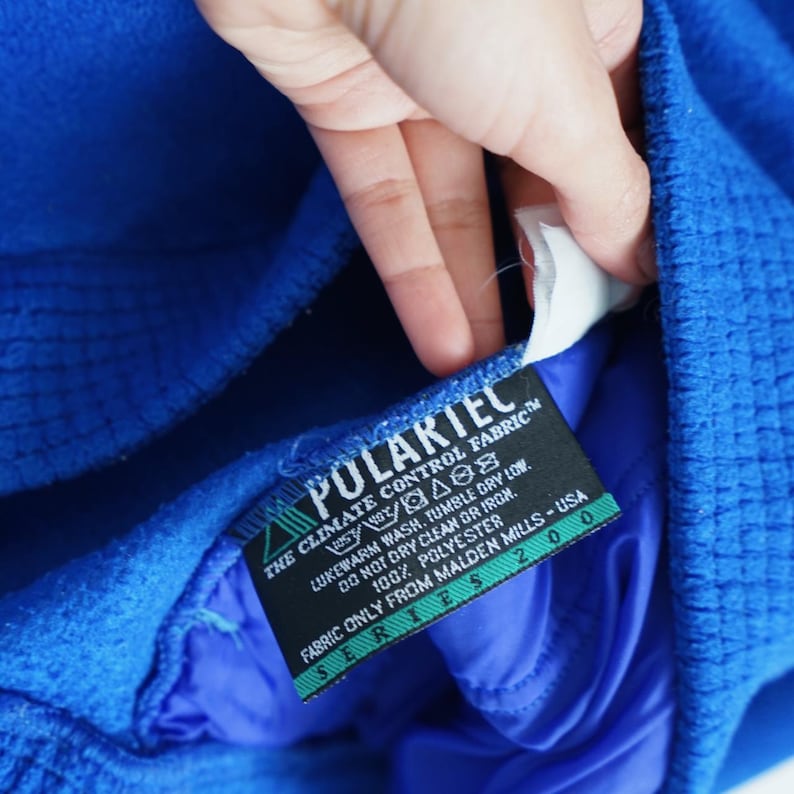 Op de afbeelding: Close-up van een felblauw kledingstuk met een getextureerd oppervlak. Een zwart label met witte tekst luidt "POLARTEC THE CLIMATE CONTROL FABRIC". Het label bevat ook wasvoorschriften en de tekst "FABRIC ONLY FROM MALDEN MILLS - USA."