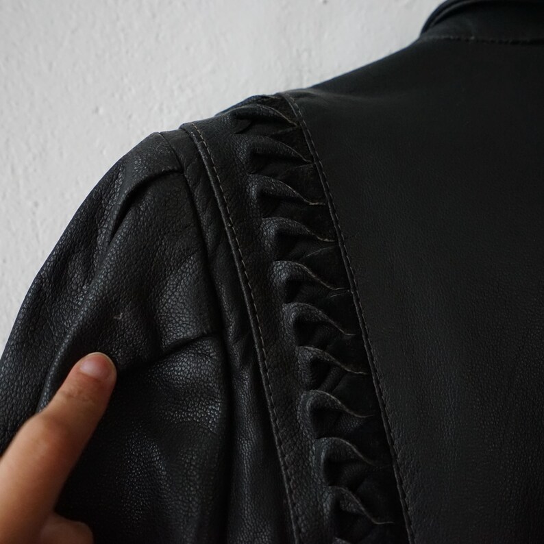May include: Close-up of a black leather jacket. The jacket features a detailed design on the shoulder and back, with a textured surface. The stitching and decorative elements are visible, highlighting the craftsmanship of the garment.