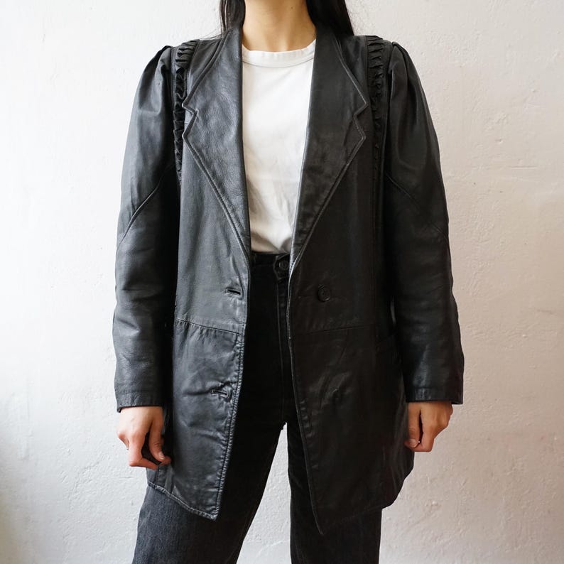 May include: A black leather blazer with a notched collar and two-button closure. The blazer features ruffled detailing on the shoulders and a tailored fit. The jacket is worn over a white t-shirt and black pants.
