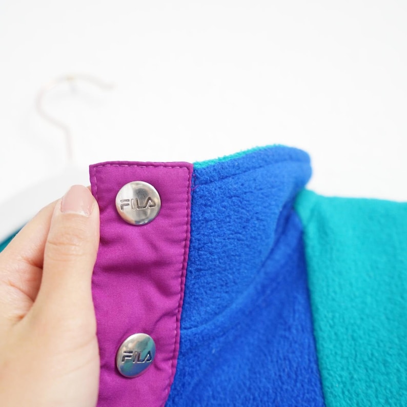 Op de afbeelding: Close-up van een kleurrijke fleece jackkraag. De jas heeft een levendig colorblock-ontwerp met blauwe, turquoise en paarse panelen. Zilveren knopen met het "FILA"-logo bevestigen de kraag. Het zachte fleecemateriaal is zichtbaar.