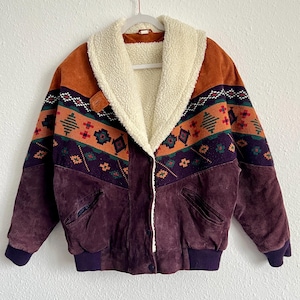 May include: A vintage suede jacket with a cream-coloured shearling collar. The jacket features a geometric pattern in orange, green, and purple across the shoulders and upper chest. The lower portion is a deep purple suede, with matching ribbed cuffs.