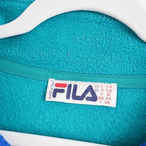 Op de afbeelding: Close-up van een turquoise fleecejack met een blauwe kraag en een witte hanger. Het Fila-logo is zichtbaar op een label, samen met maat informatie. De jas is gemaakt van een zacht, getextureerd materiaal.