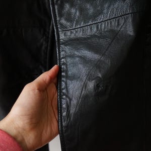 May include: Close-up of a black leather jacket. The jacket's texture is visible, with seams and subtle variations in the leather's surface. The jacket appears to be well-worn, with a classic design.