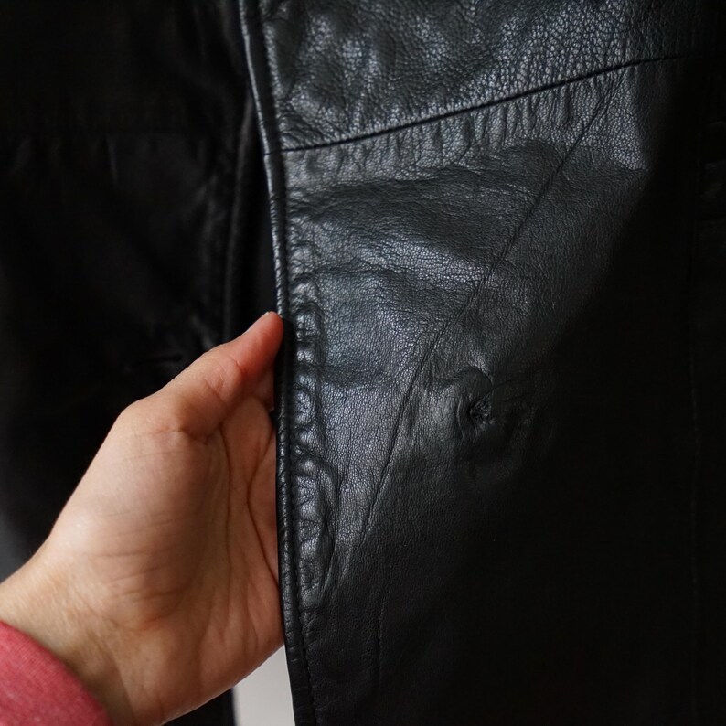 May include: Close-up of a black leather jacket. The jacket's texture is visible, with seams and subtle variations in the leather's surface. The jacket appears to be well-worn, with a classic design.