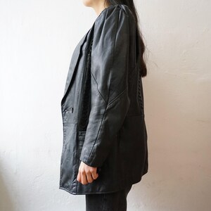 May include: Black leather blazer with a notched lapel and two-button closure. The jacket has padded shoulders and a tailored fit. The sleeves are long, and the blazer falls to mid-thigh. The leather appears to be slightly distressed.
