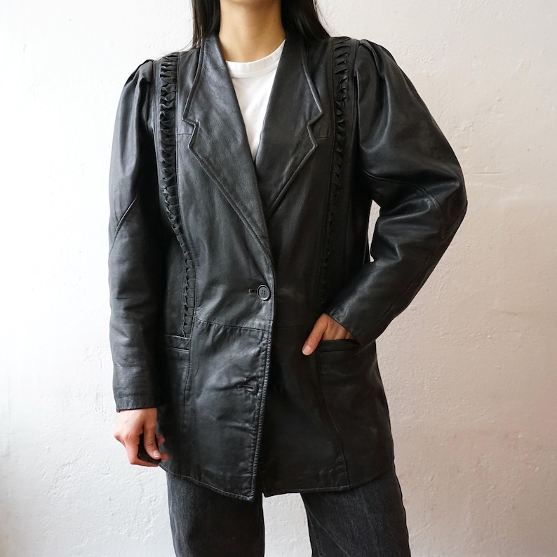 May include: Black leather blazer with shoulder detailing and a single-button closure. The blazer has a classic lapel and two front pockets. The sleeves have a slight puff at the shoulder. The jacket is worn over a white t-shirt and dark jeans.