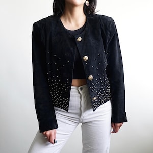 Könnte beinhalten: Eine schwarze Wildlederjacke mit rundem Ausschnitt, langen Ärmeln und vier großen goldfarbenen Metallknöpfen auf der Vorderseite. Der untere Teil der Jacke ist mit einem dichten Muster aus kleinen silberfarbenen Nieten verziert, die nach oben hin auslaufen.