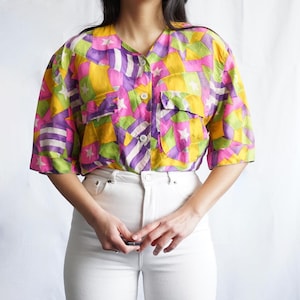 Könnte beinhalten: Ein kurzärmeliges Hemd mit Knopfleiste und einem lebendigen geometrischen Muster in Gelb, Rosa, Lila, Grün und Weiß. Das Hemd hat einen V-Ausschnitt, zwei Brusttaschen und weiße Knöpfe. Es wird mit einer weißen Jeans kombiniert.