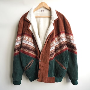 May include: A brown and dark green jacket featuring a white sherpa lining and collar. The garment displays a horizontal band with geometric patterns in white, brown, and black across the chest and sleeves, with two front pockets.
