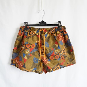 Könnte beinhalten: Shorts mit olivgrüner Grundfarbe und einem geometrischen Muster in Rot, Blau und Schwarz. Die Shorts haben einen elastischen Bund mit orangefarbenem Kordelzug und hängen an einem schwarzen Kunststoffbügel vor einem schlichten weißen Hintergrund.