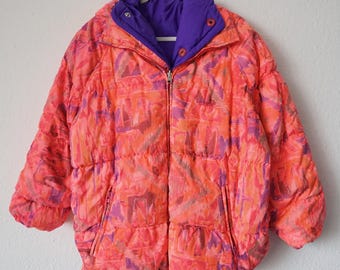May include: Reversible puffer jacket with an abstract pattern in orange, pink, and purple. It has a zip closure and a purple interior. The collar and cuffs are also purple. The jacket is hanging on a white hanger.