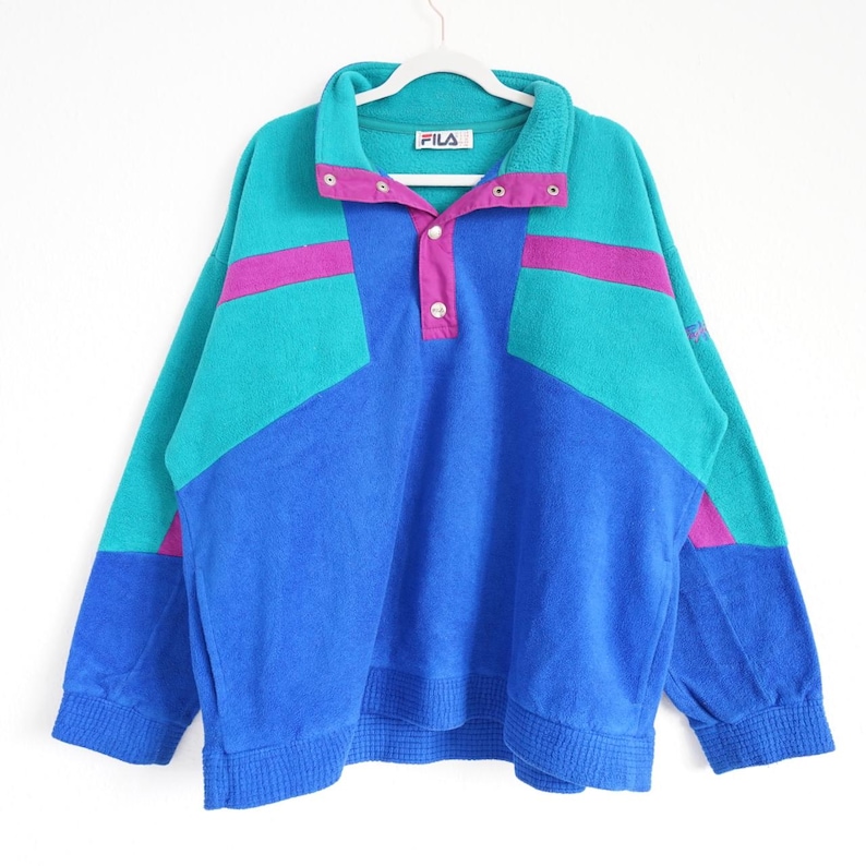 Op de afbeelding: Een vintage Fila sweatshirt in tinten blauw, teal en magenta. De top met lange mouwen heeft een kraag met drukknoopsluitingen en een colorblock-ontwerp. Het Fila-logo is zichtbaar op de kraag.