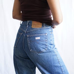 Könnte beinhalten: Hoch taillierte blaue Jeans aus Denim mit klassischer Passform. Die Jeans haben eine Gesäßtasche mit kleinem Logo und einen Lederaufnäher am Bund. Das Model trägt ein braunes T-Shirt.