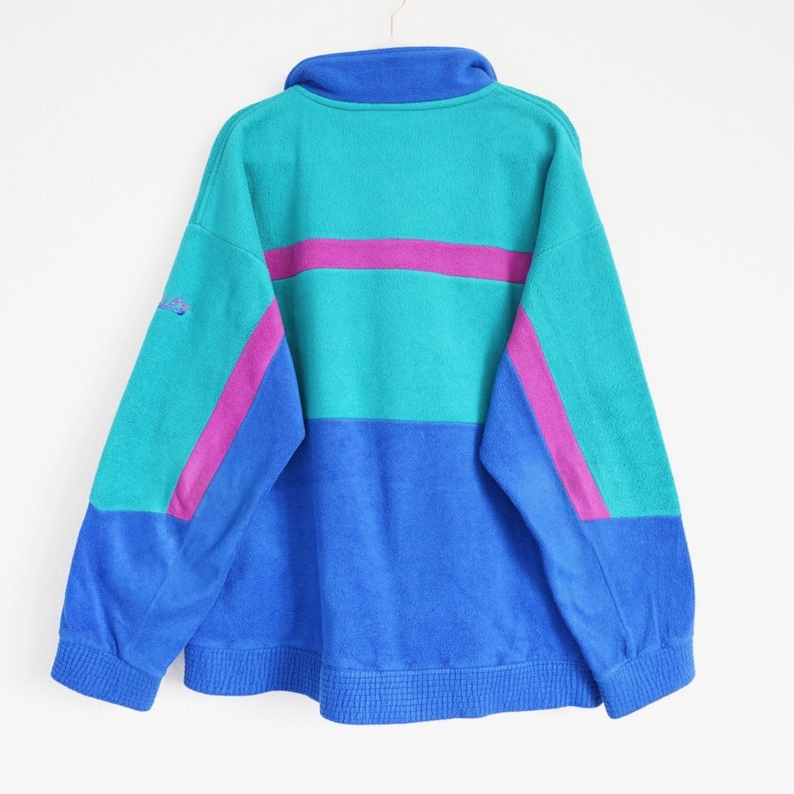 Op de afbeelding: Een vintage fleece trui met kleurblokken in blauw, teal en magenta tinten. De retro-stijl sweatshirt heeft een hoge kraag en een horizontale magenta streep over de borst. De mouwen hebben magenta strepen. Een klein logo is zichtbaar op de linkermouw.