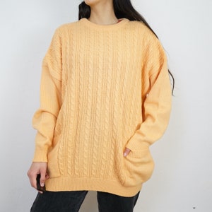 Könnte beinhalten: Ein hellorangefarbener Zopfstrickpullover mit Rundhalsausschnitt und zwei Vordertaschen. Der Pullover hat eine lockere Passform und lange Ärmel. Das Zopfmuster ist auf der Vorderseite und den Ärmeln sichtbar. Der Pullover ist aus weichem, bequemem Material.