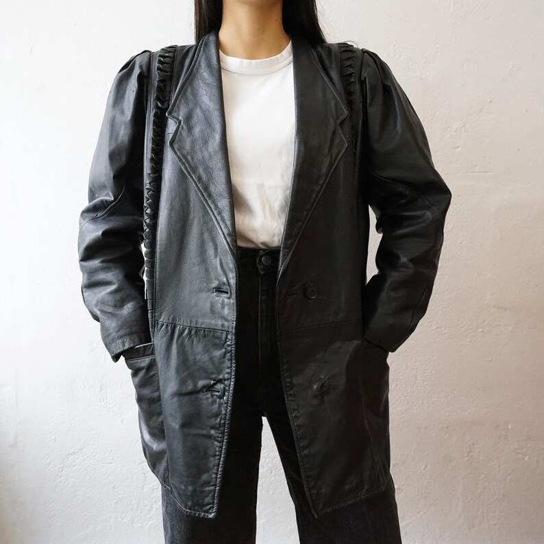 May include: A black leather blazer with a notched collar, two front pockets, and a double-breasted closure. The blazer has padded shoulders and a longline silhouette. The sleeves have a unique braided detail.