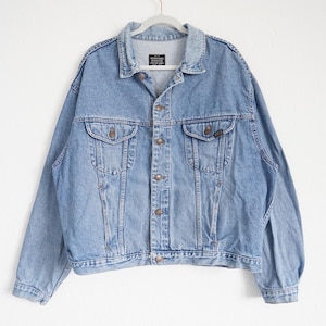 Könnte beinhalten: Hellblaue Jeansjacke mit Knopfleiste, zwei Brusttaschen und zwei Seitentaschen. Die Jacke hat einen klassischen Kragen und lange Ärmel. Die Jacke hängt an einem weißen Kleiderbügel vor weißem Hintergrund.