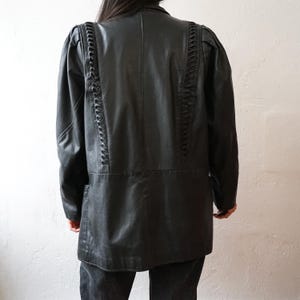 May include: Black leather jacket with decorative braided detailing along the shoulders and back. The jacket has a classic silhouette with a tailored fit, long sleeves, and a button-down front. The jacket is a versatile piece for any wardrobe.
