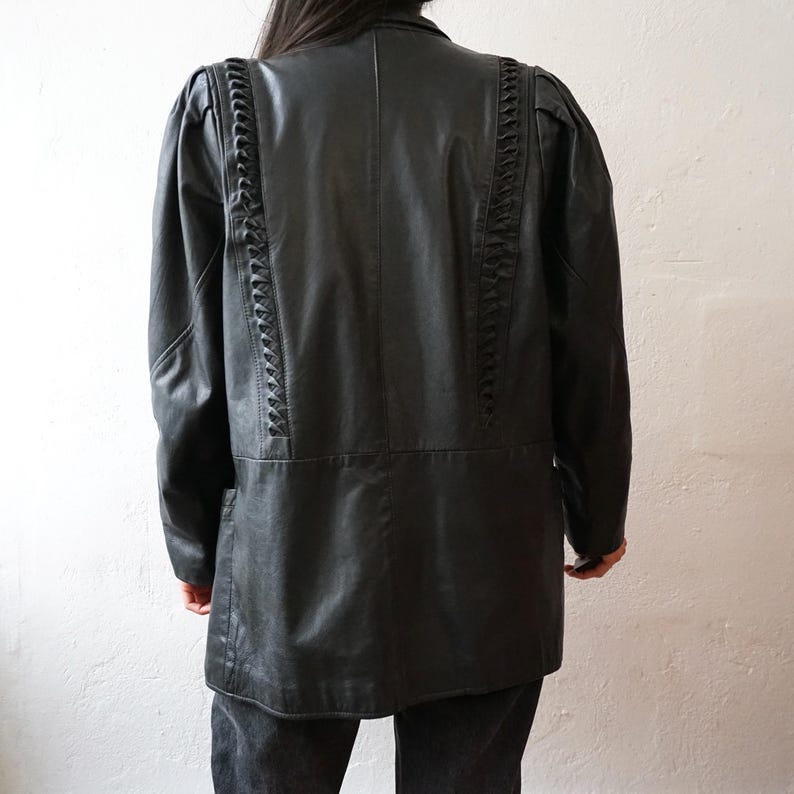 May include: Black leather jacket with decorative braided detailing along the shoulders and back. The jacket has a classic silhouette with a tailored fit, long sleeves, and a button-down front. The jacket is a versatile piece for any wardrobe.