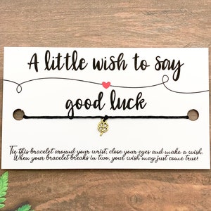 May include: A black string bracelet with a gold four-leaf clover charm, presented on a white card. The card reads "A little wish to say good luck" with additional text about making a wish.