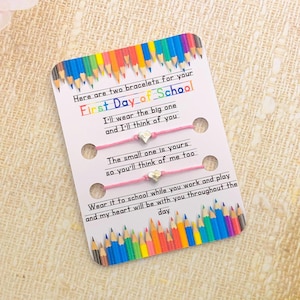 May include: Two pink friendship bracelets with silver heart charms. The card reads "Here are two bracelets for your First Day of School. I'll wear the big one and I'll think of you. The small one is yours so you'll think of me too. Wear it to school while you work and play and my heart will be with you throughout the day."