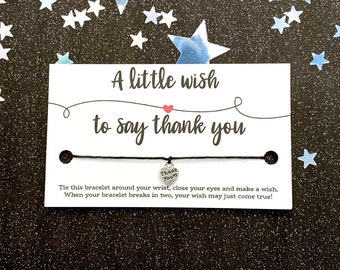 May include: A white card with the text "A little wish to say thank you" and a small heart. A black string bracelet with a silver heart charm that says "Thank You". The background is black with blue and white star confetti.