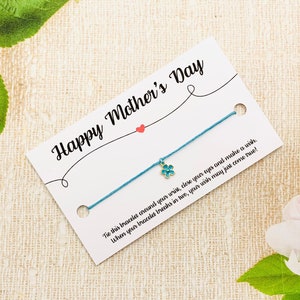 Puede incluir: Un brazalete de amistad azul con un pequeño dije de flor azul y una tarjeta blanca que dice "Feliz Día de la Madre". Ata este brazalete alrededor de tu muñeca, cierra los ojos y pide un deseo. Cuando tu brazalete se rompa en dos, tu deseo puede hacerse realidad!