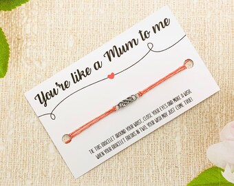 May include: A coral-colored string bracelet with a silver-toned infinity charm, presented on a white card. The card reads "You're like a Mum to me" with a small red heart. Text below instructs the wearer to make a wish.