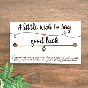 Puede incluir: Una pulsera de la buena suerte en una tarjeta blanca con el texto "A little wish to say good luck". La pulsera es de color verde oliva con un dije de trébol de cuatro hojas plateado. La tarjeta incluye instrucciones.