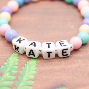 May include: A pastel-colored beaded bracelet with white letter beads spelling out the name "KATE".