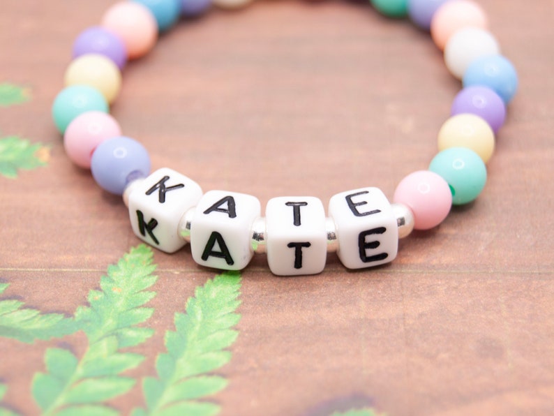 May include: A pastel-colored beaded bracelet with white letter beads spelling out the name "KATE".