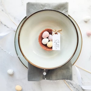 May include: A white ceramic bowl with a brown rim sits on a grey napkin. A small nest with pastel coloured Easter eggs is inside the bowl. A white card with the word "Stelle" is attached to the nest.