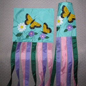 May include: A turquoise windsock with yellow butterflies and purple flowers. The windsock has multiple streamers in pink, purple, and green.