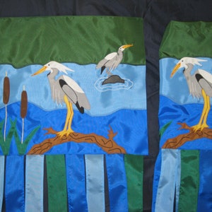 May include: Two blue and green fabric wall hangings with embroidered images of gray herons standing on branches over blue water. The hangings have blue, green, and white fabric fringe.