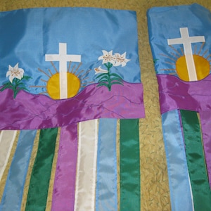 May include: Two blue and purple fabric banners with a white cross, a yellow sun, and white lilies. The banners have multiple purple, white, green, and blue streamers hanging from the bottom.