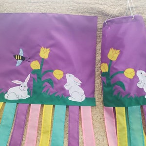 May include: Two purple fabric banners with white bunnies, yellow tulips, and green leaves. The banners have a fringe of purple, pink, yellow, and blue ribbons.
