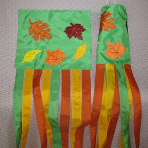 May include: A green fabric windsock with orange, yellow, and brown leaf designs. The windsock has multiple green, orange, and yellow streamers.