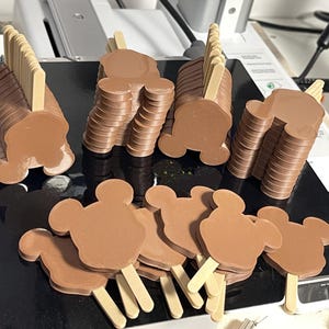 May include: Brown, Mickey Mouse-shaped ice cream bars on wooden sticks, stacked in several piles. The ice cream bars are arranged on a black surface, with some individual bars scattered around. The image is taken from a high angle, showing the tops and sides of the bars.