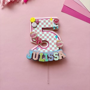 Peut inclure: Un cake topper numéro cinq à carreaux roses et blancs avec le nom "JULISSA" en lettres colorées. Le topper est décoré de visages souriants, de cœurs, d'un patin à roulettes et d'accents de boule disco. Un bâtonnet en plastique transparent soutient le topper.