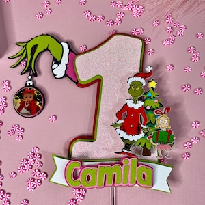 May include: A festive cake topper featuring a Grinch theme. The design includes a large number one, the Grinch, a Christmas tree, and the name "Camila" in green and white. A small photo is in a Christmas ornament.