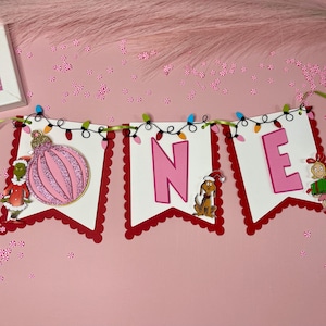 May include: A festive banner spelling "ONE" with pink letters and a decorative ornament. The banner is adorned with Christmas lights and Grinch-themed characters, perfect for a birthday celebration.