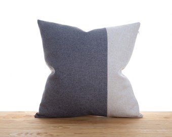 Puede incluir: Un cojín decorativo cuadrado con un diseño bicolor. Una mitad es gris azulado oscuro y la otra gris claro. El cojín está hecho de tela texturizada y está sobre una superficie de madera.
